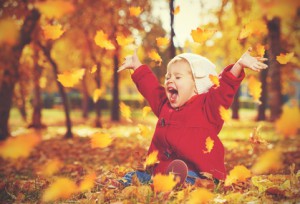 kleines Kind, im Herbstlaub sitzend streckt die Hände in die Luft und schreit vor Freude